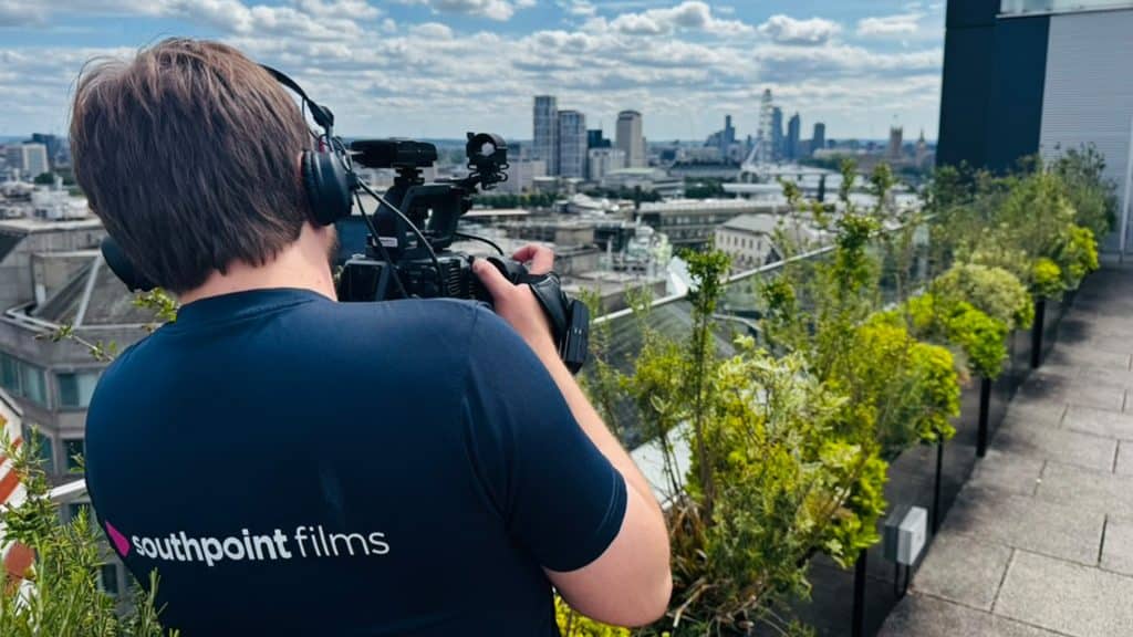 A person with light brown hair wearing a navy blue "southpoint films" t-shirt and headphones films the London skyline with a professional video camera from a rooftop garden.