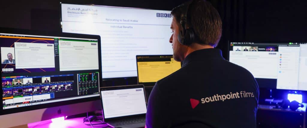A person wearing a "southpoint films" polo shirt and headphones monitors multiple screens displaying live video production software and a digital presentation.