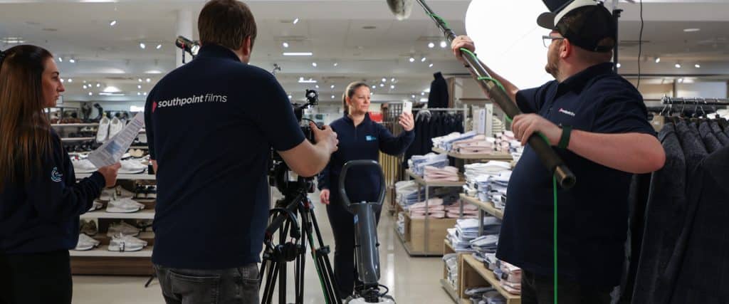 A film crew wearing Southpoint Films apparel operates a camera, lighting, and audio equipment while filming in a retail clothing store.
