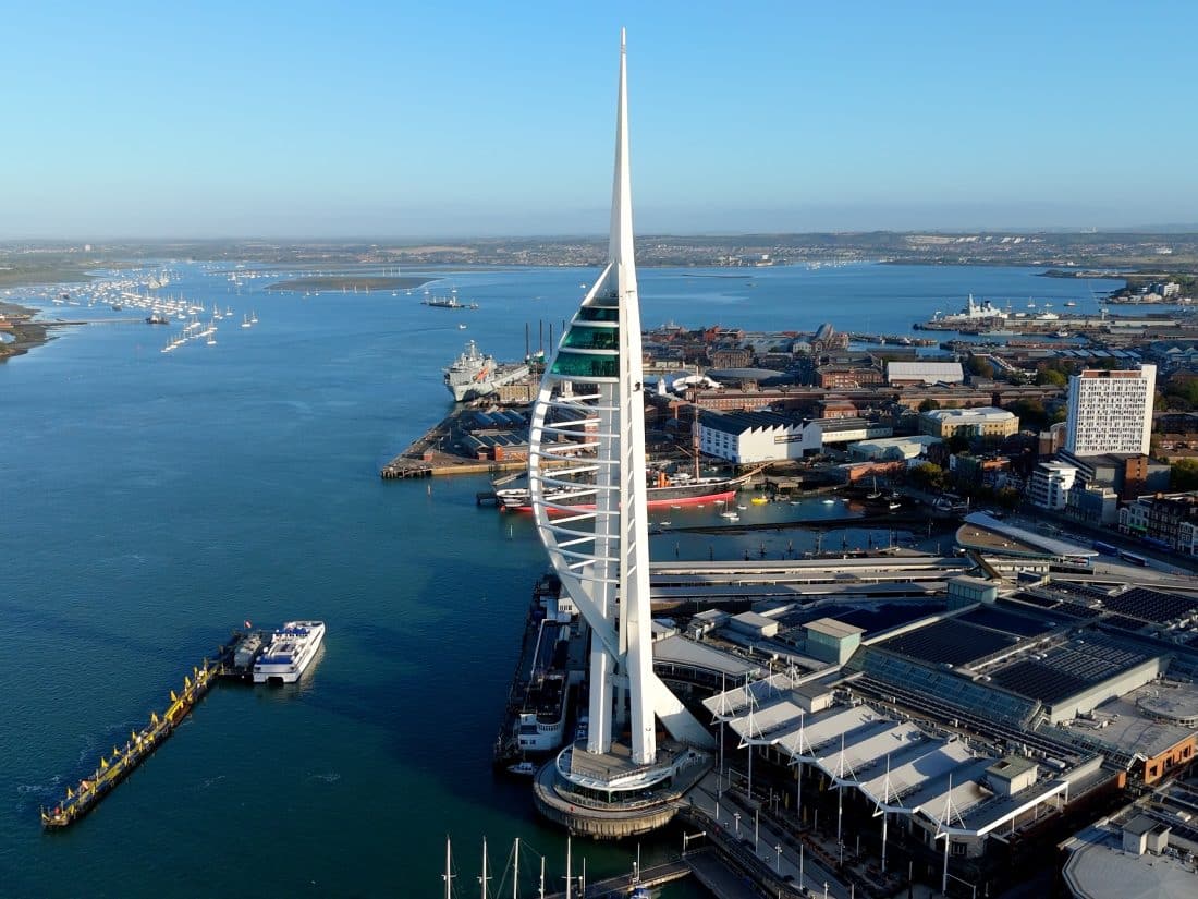 An aerial view of the white, sail-shaped Spinnaker Tower overlooking Portsmouth Harbour and surrounding city buildings under a clear blue sky.