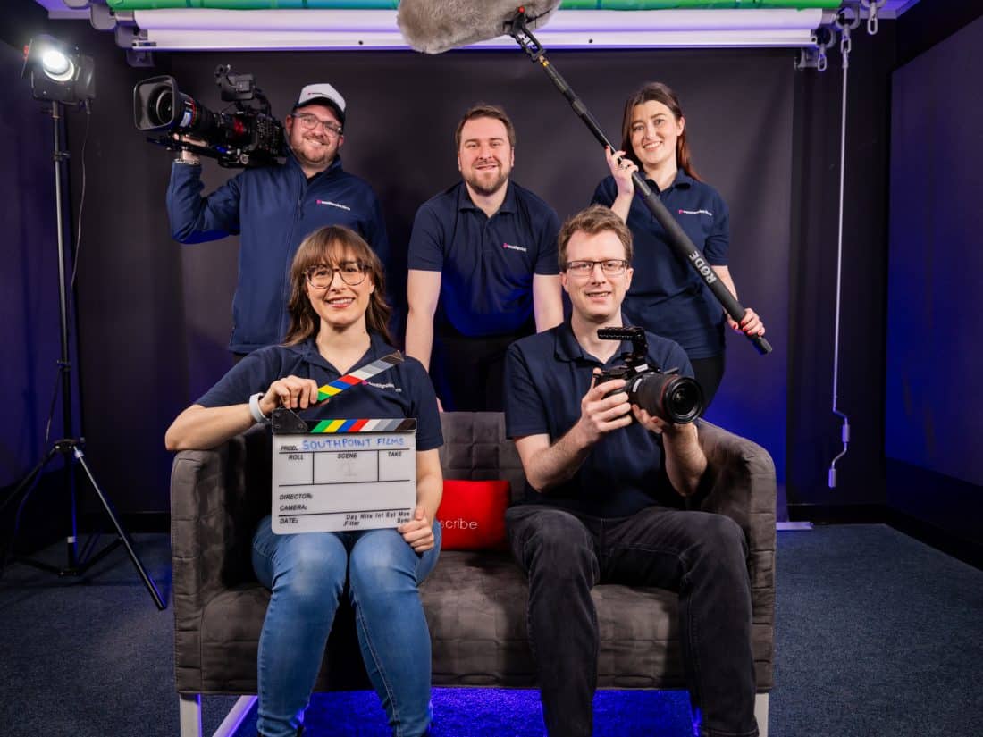 Five team members wearing navy blue branded clothing pose in a film studio with professional equipment including cameras, a boom microphone, and a clapperboard.