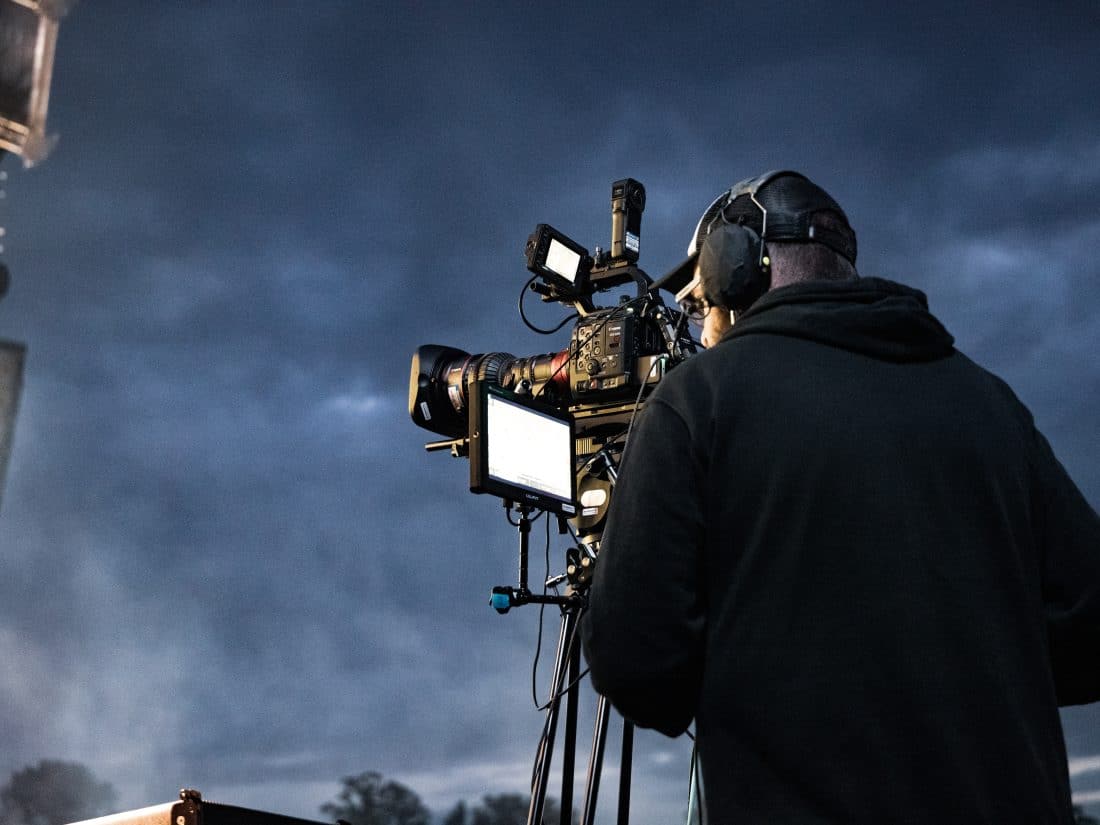 A camera operator in a dark hoodie and headset operates a professional cinema camera on a tripod against a cloudy, twilight sky.