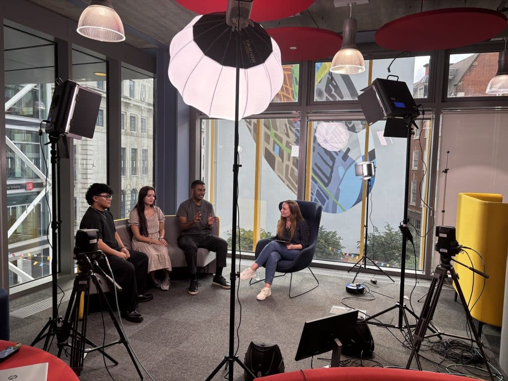 Four people sit in a professional studio setting for an interview, with two cameras on tripods and several large studio lights positioned around them. A person in a blue chair holds a tablet while facing three individuals seated on a grey sofa against a backdrop of large windows.