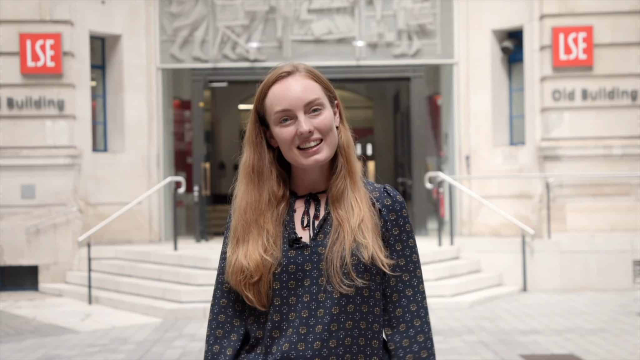 A person with long strawberry-blonde hair and a patterned navy blouse stands smiling in front of the London School of Economics (LSE) Old Building entrance. The background features stone architecture, red LSE logos, and a set of wide steps leading to glass doors.