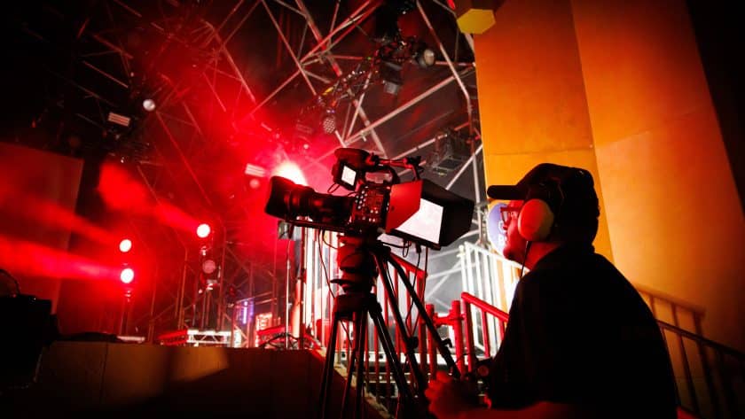 A camera operator wearing a black cap and noise-canceling headphones films a live event stage bathed in intense red lighting and hazy atmosphere. The professional video camera is mounted on a tripod in the foreground, capturing the industrial scaffolding and bright spotlights of the performance area.