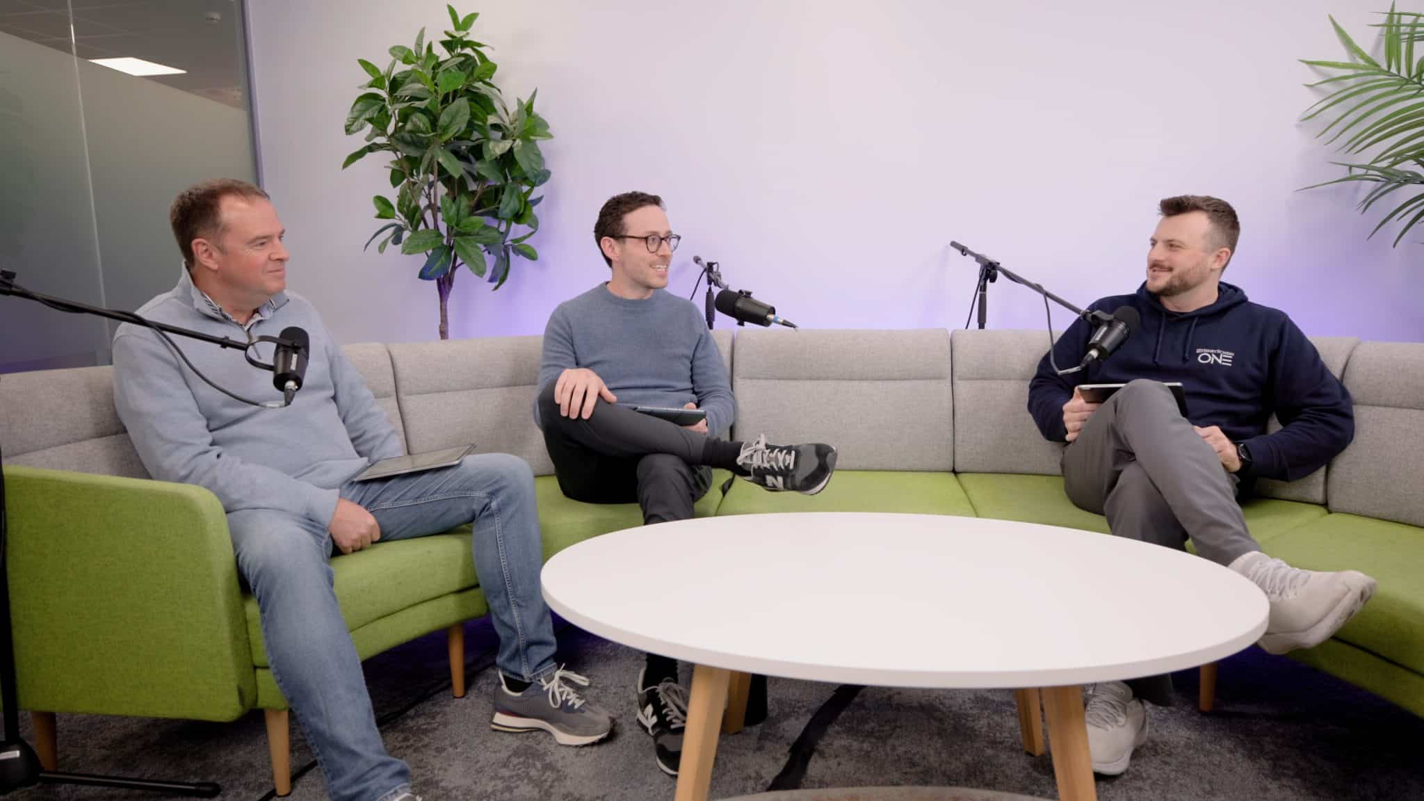 Three men sit on a modern lime green and gray curved sofa in a brightly lit studio, engaged in a conversation with professional microphones positioned in front of each person. The setting includes a round white coffee table in the foreground and indoor plants in the background.
