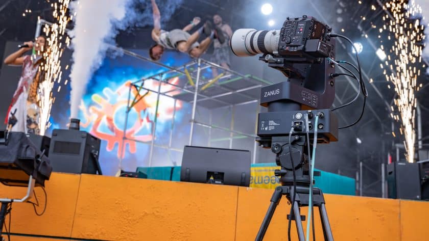 A professional video camera mounted on a robotic ZANUS tripod head captures a high-energy stage performance featuring a singer, pyrotechnic sparks, and an acrobat mid-flip on a metal scaffolding structure. The background includes a large vibrant digital screen and bright stage lighting.