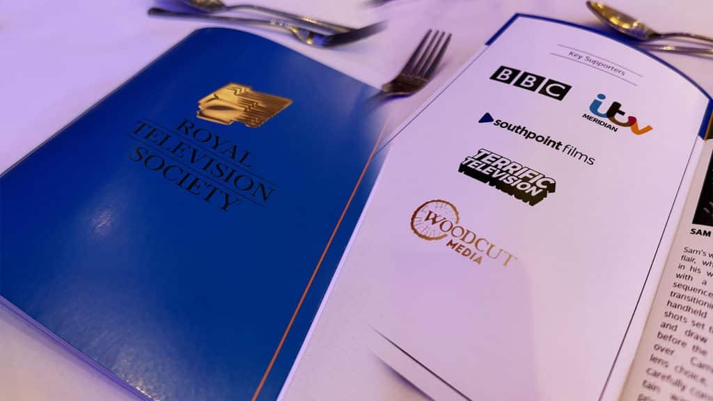 A blue Royal Television Society program rests on a white tablecloth next to an open page listing key supporters, including logos for the BBC, ITV Meridian, Southpoint Films, Terrific Television, and Woodcut Media. Silver cutlery is visible in the blurred background.