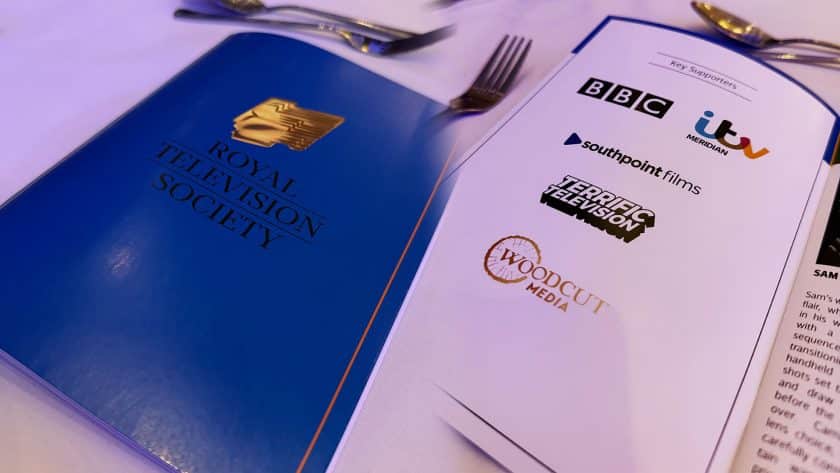 A blue Royal Television Society program rests on a white tablecloth next to an open page listing key supporters, including logos for the BBC, ITV Meridian, Southpoint Films, Terrific Television, and Woodcut Media. Silver cutlery is visible in the blurred background.