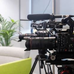 A professional Canon EOS C400 cinema camera with a large lens and a Rode shotgun microphone is mounted on a tripod in a bright studio. In the blurred background, a second camera setup and a green potted plant are visible next to a modern sofa.