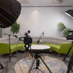 SPF 2026aProfessional video production studio setup featuring four cameras on tripods, overhead softbox lights, and microphones arranged around a circular lime green and grey sofa and a white coffee table. A round patterned rug sits on the floor in the centre of the recording area.