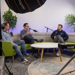Three people sit on a curved green and grey sofa in a professional studio, engaged in a recorded conversation with microphones and cameras visible in the foreground. Large studio lights and potted plants frame the set, which includes a round white coffee table on a patterned rug.