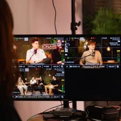 A person viewed from behind monitors a live broadcast on a screen displaying multiple camera feeds of individuals in a professional podcast studio setting. The screen shows technical data overlays, an "ON AIR" sign, and people seated with microphones in a warmly lit environment with acoustic paneling and plants.