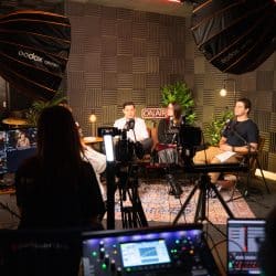 Three people sit on a patterned rug in a professional podcast studio, surrounded by soundproofing foam, professional lighting, and cameras. In the foreground, a production crew member monitors the recording on a multi-view screen and audio mixer.