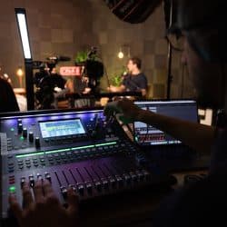 An audio engineer operates an Allen & Heath digital mixing console in the foreground, while a person sits for an interview under studio lights in the background. A laptop displays audio waveforms next to the mixing board in a professional recording environment.