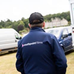 A person wearing a navy blue "southpoint films" jacket and a black mesh cap looks toward a large manor house in the distance.