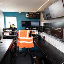 The interior of a mobile production trailer features multiple monitors displaying video feeds, professional audio speakers, and an orange high-visibility vest draped over a chair.