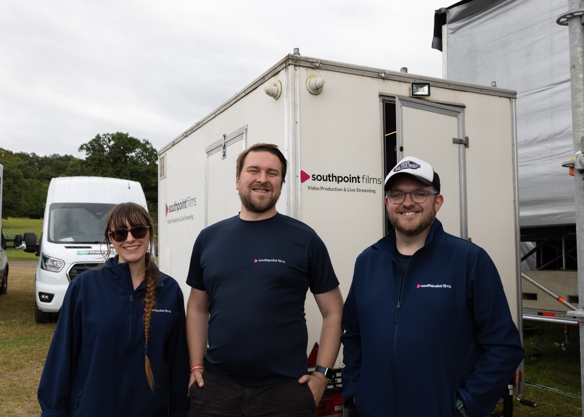Three people wearing navy blue "southpoint films" apparel stand smiling in front of a white mobile production trailer.