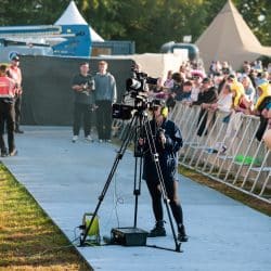 A camera operator stands behind a large broadcast camera on a tripod, filming a crowd of people at an outdoor event.