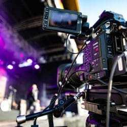 A professional Canon cinema camera is mounted on a tripod in front of an outdoor festival stage with lighting rigs.