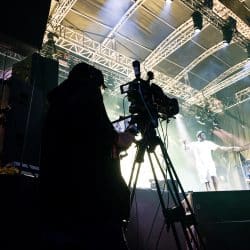 A silhouette of a camera operator filming a live musical performance on a brightly lit stage under a metal truss system.