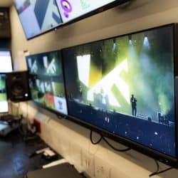 A row of computer monitors inside a production trailer displays a live video feed of a musical performer on a stage with bright yellow lighting.