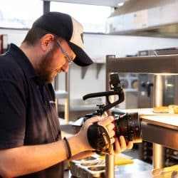 A person wearing a black and white baseball cap and a polo shirt films food on a stainless steel kitchen shelf using a professional video camera.