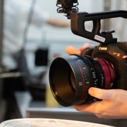 A close-up shot of a person’s hands holding a professional Canon video camera with a large cinema lens, focused on a plate in the foreground.