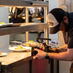 A person with a beard and a baseball cap leans over a stainless steel counter to film plated dishes with a professional camera in a commercial kitchen.