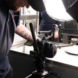 A person with a tattoo on their forearm operates a professional camera mounted on a tripod, aimed at a white plate on a kitchen counter under bright studio lighting.