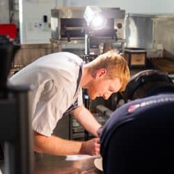 A chef with red hair and a white uniform leans over a counter to plate food while being filmed by a person in a dark polo shirt under a bright studio light.