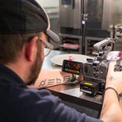 A high-angle view from behind a person showing their arm tattoos and a professional camera as they film food on a plate in a kitchen setting.