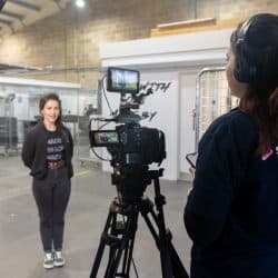 A person wearing headphones and a navy polo shirt operates a professional video camera on a tripod, filming a person standing in a warehouse-style space.