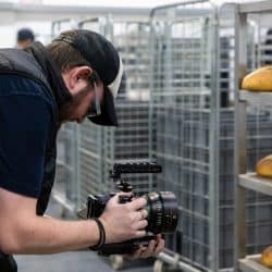 A person wearing a black quilted vest and a baseball cap holds a professional cinema camera, focusing on several loaves of fresh bread on a metal rack.