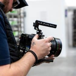 A close-up of a person's hands holding a professional camera with an 85mm lens; a small star tattoo is visible on the person's hand.