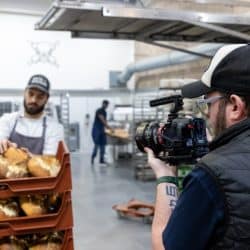 A person films a baker in a white uniform and grey apron as they organize fresh loaves of bread on a red plastic tray in a bakery.