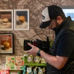 A person wearing a black vest and a baseball cap holds a professional video camera while standing in front of a wall decorated with coffee-themed posters and a map.