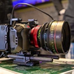 A close-up shot of a professional camera rig with a large 85mm cinema lens resting on a table over some printed documents.