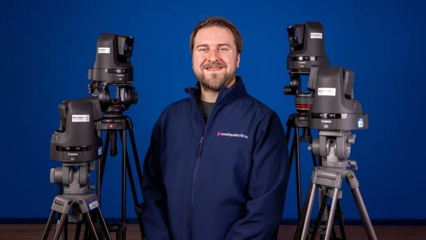 A smiling person wearing a dark blue jacket featuring the "Southpoint Films" logo stands in front of a blue backdrop, surrounded by four Canon PTZ cameras mounted on tripods.